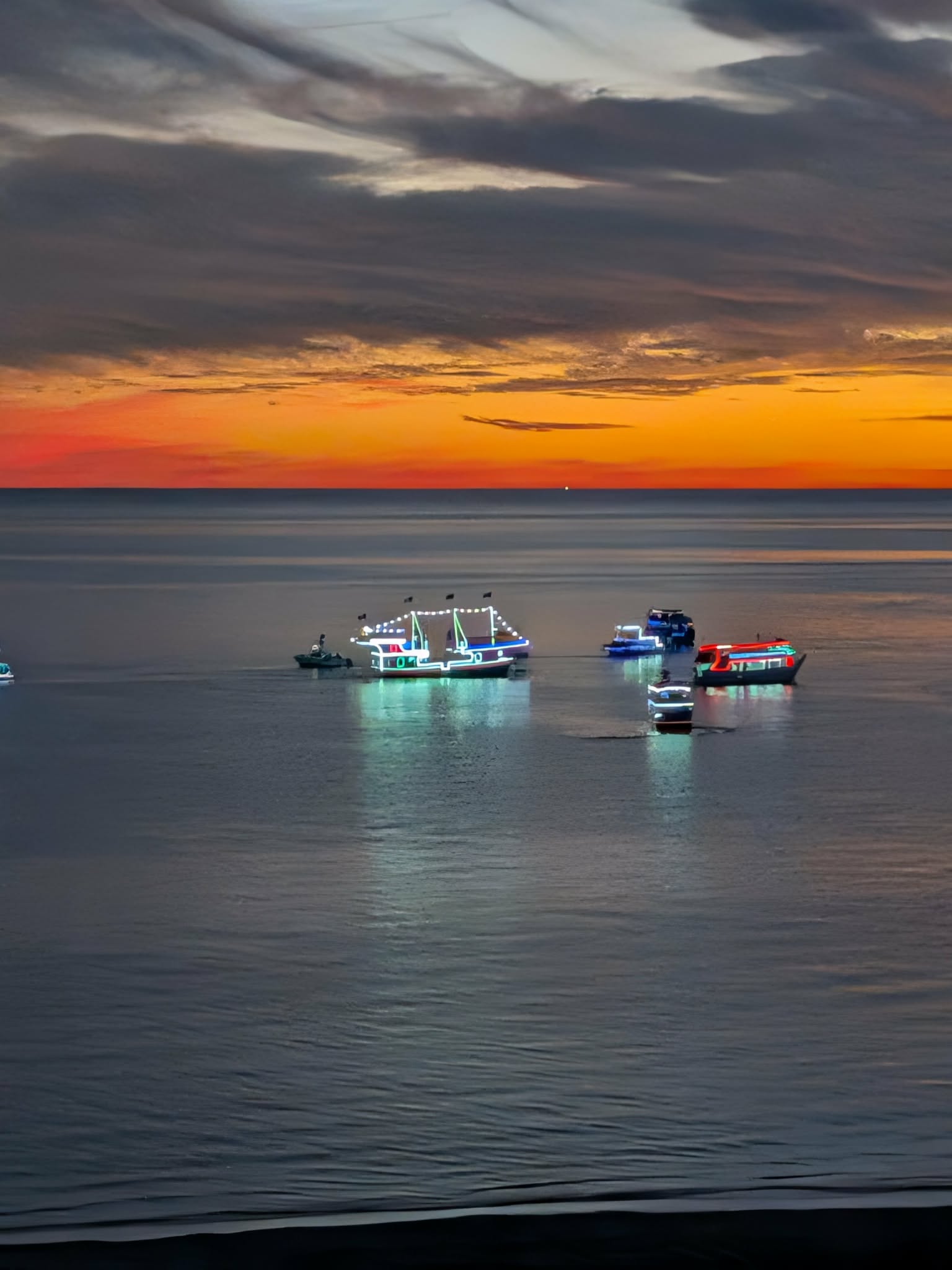 Boats outlined in lights on calm water beneath an orange sunset sky